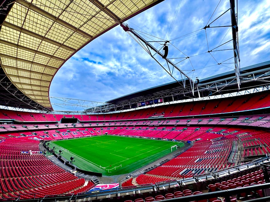 Interior view of Wembley Arena stadium featuring a well-maintained grass football pitch at the centre, surrounded by tiered red seating sections. The arena's curved roof structure and steel support be
